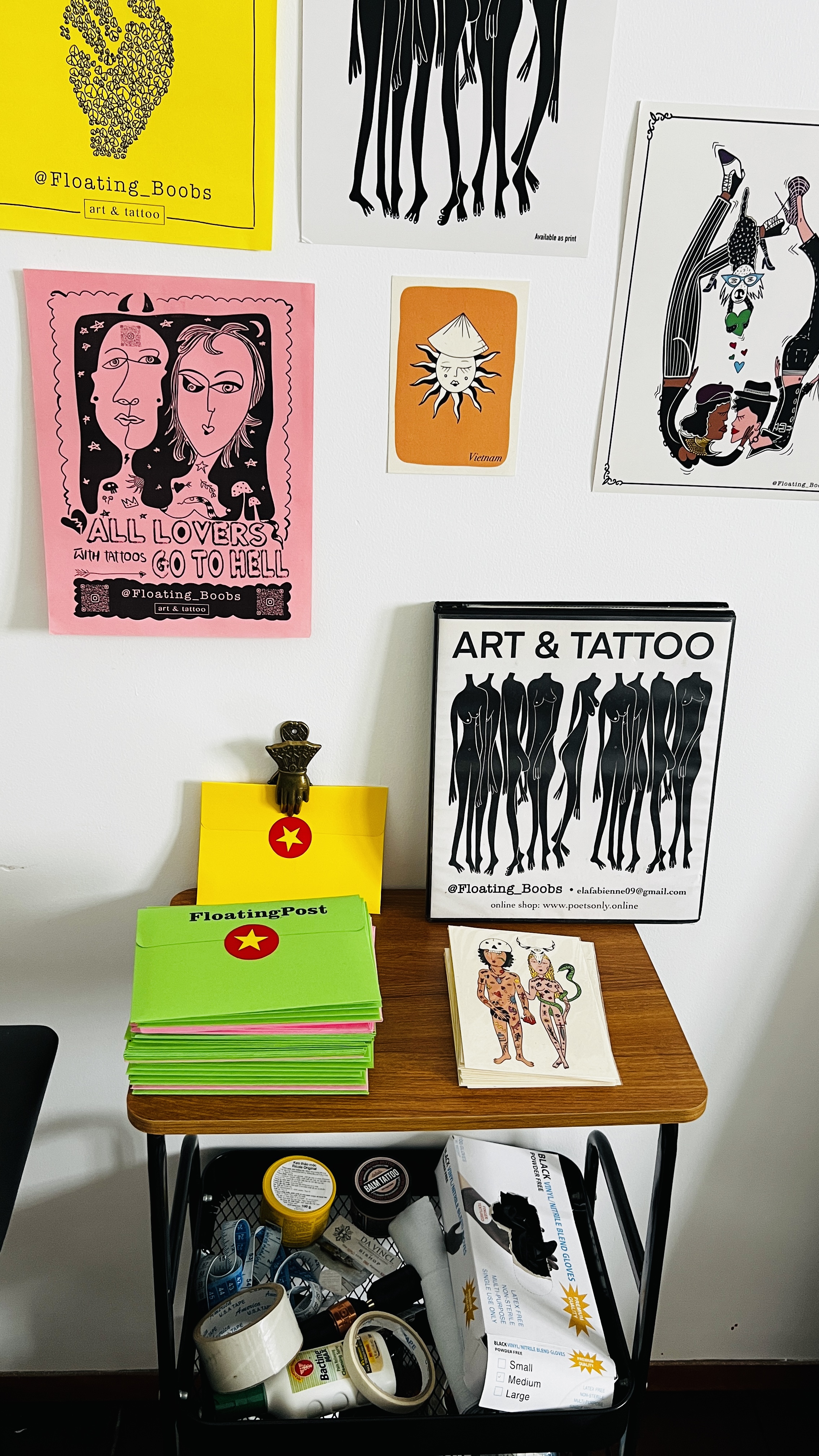 Ela's Hội An studio wall covered in Floating Boobs art prints, with a stack of FloatingPost envelopes and the Art & Tattoo portfolio on the desk below