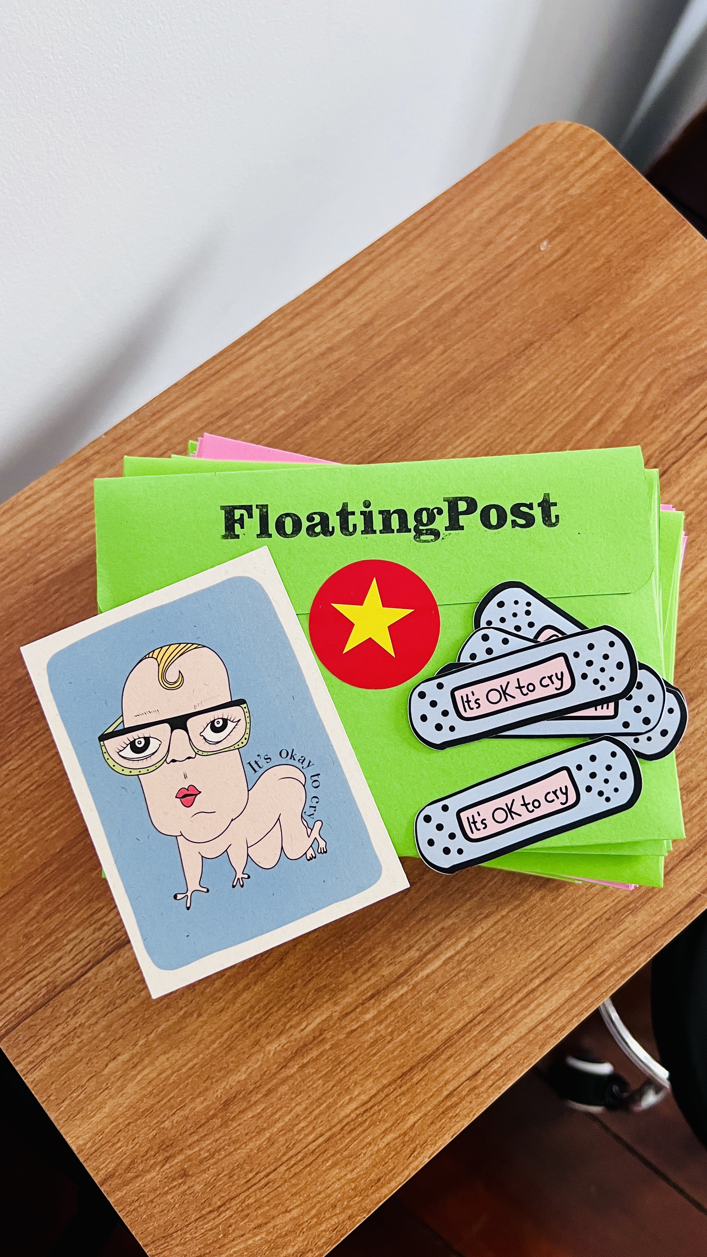 An It's OK to cry art card and bandaid stickers on top of a stack of green Floating Post envelopes